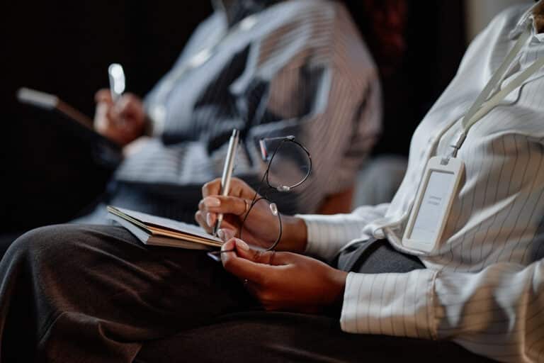 Close up on Black female conference participant holding glasses in hand while keeping notes on speakers presentation writing in notebook during business meeting in auditorium, copy space. Close up on Black female conference participant holding glasses in hand while keeping notes on speakers presentation writing in notebook during business meeting in auditorium, copy space. Competitive Evaluation
