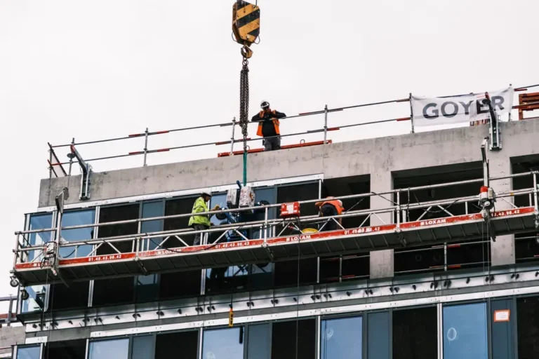 The image shows construction workers installing a large glass panel on a high-rise building exterior using a crane and a suspended staging platform.