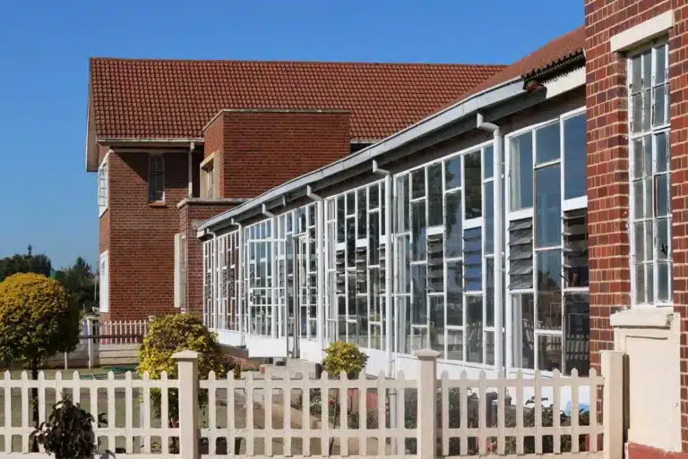 The image shows an exterior view of a red-brick school or institutional building featuring an attached sunroom or glass-enclosed corridor with large multi-pane white window frames and a red-tiled roof.