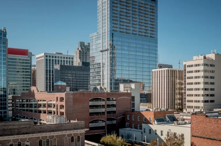 The image shows the skyline of downtown Raleigh, North Carolina, featuring urban high-rises and historic brick buildings.