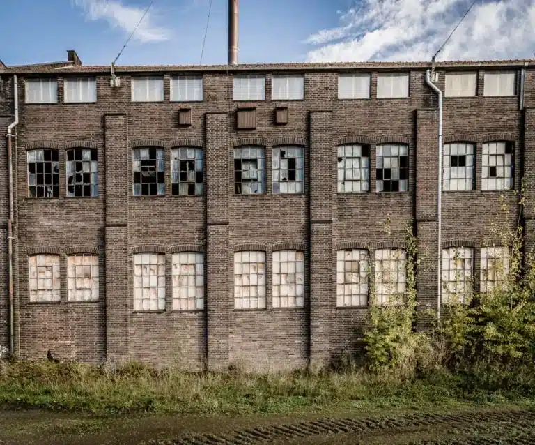 The image shows the brick facade of an old, abandoned industrial factory building featuring weathered brick walls, multi-pane steel-framed windows with several broken or boarded-up panes, and sparse vegetation along the ground.